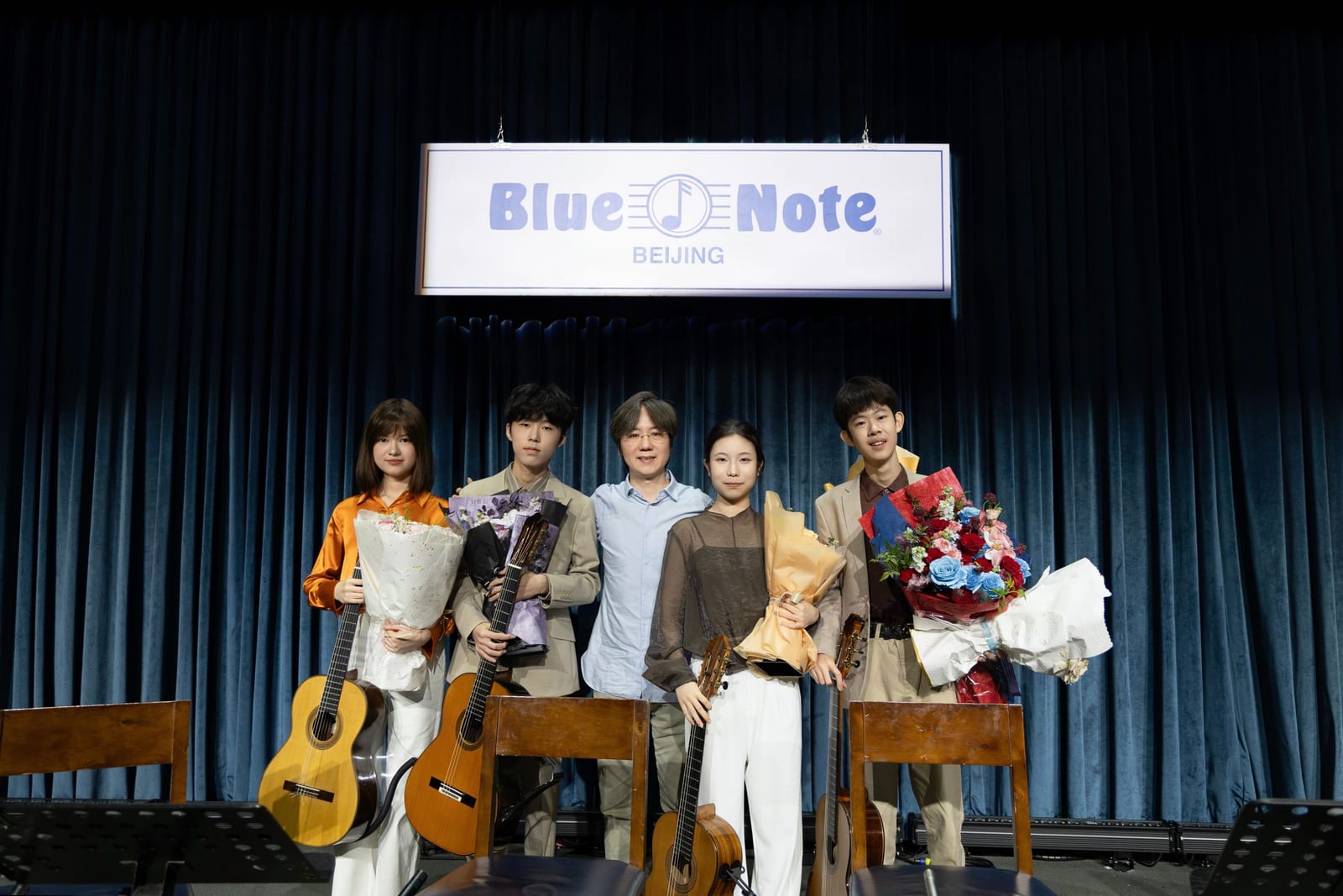 After the concert, five people stand together under the Blue Note Beijing sign; the four players hold their guitars and flowers.